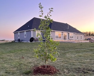A single-story house with white siding and black roof during sunset. A young tree in the foreground with a fresh mulch circle around it.