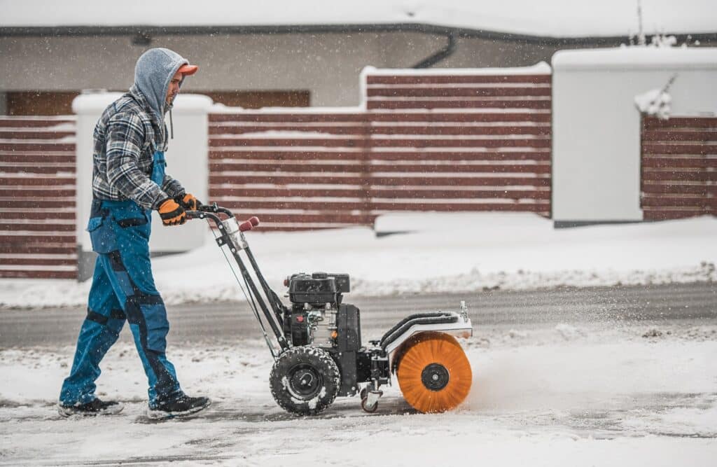 A person is operating a snowblower on a snowy pavement. They're wearing winter clothing, including a hood, gloves, and boots, against a snowy backdrop.