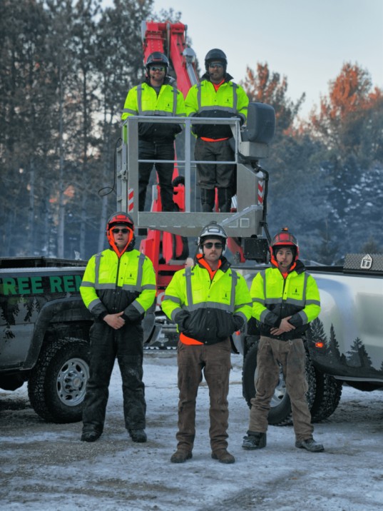 The RauschTree team in Wisconsin, wearing bright yellow safety gear, stands together with a tree removal truck and lift equipment in the background, ready for tree service operations in a snowy forest setting.