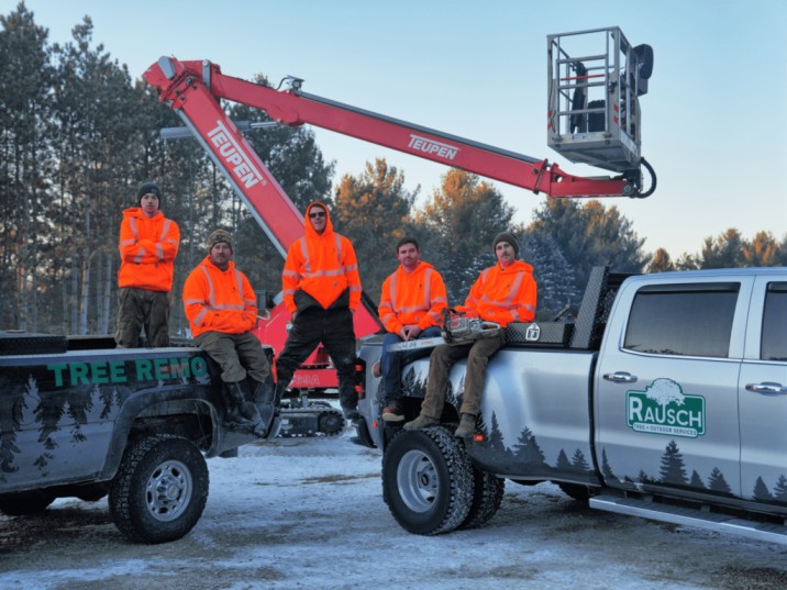 Rausch Tree Service team in Wisconsin posing in front of work trucks and a red TEUPEN lift during winter. Crew members wear bright orange safety jackets, standing and sitting on branded vehicles marked 'Tree Removal' and 'Rausch Tree & Outdoor Services.' Snow-covered ground and pine trees in the background.